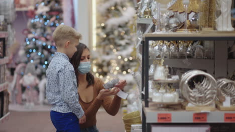 In-a-pandemic-the-coronavirus-epidemic.-A-mother-in-a-medical-mask-with-her-son-at-the-mall-choose-home-decorations-for-Christmas.-Watch-Christmas-tree-toys.-Coronavirus-epidemic